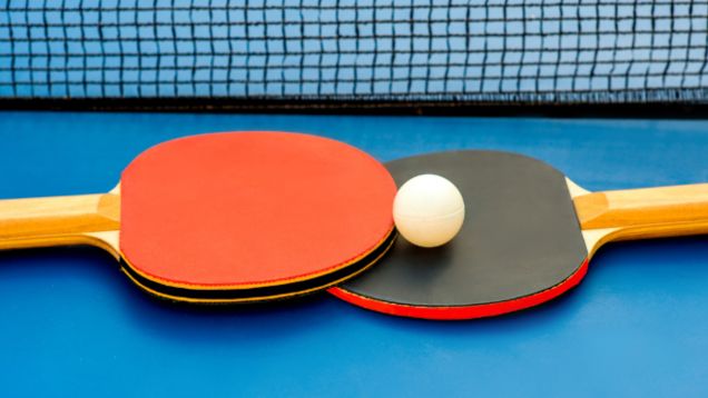 Foto in Großaufnahme einer roten und schwarzen Tischtenniskelle mit Tischtennisball auf einer blauen Tischtennisplatte